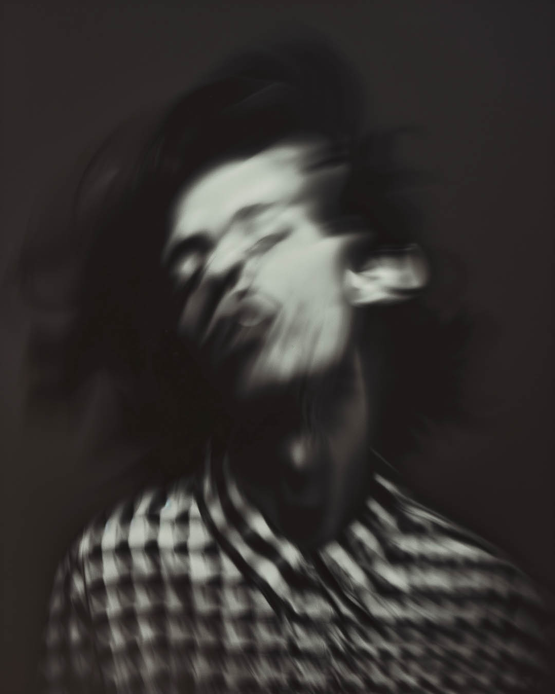 Blurred, motion-filled portrait of a man in a checkered shirt, with his head in motion creating a distorted, surreal effect.